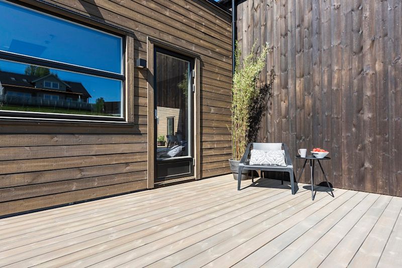 Solbelyst träfasad med ljus trädäck, svart stol med kudde och litet bord med mugg och jordgubbar.