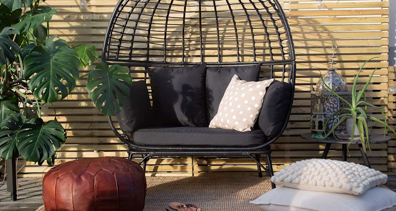 Uteplats med svart rottingsoffa, beige prickkudde, röd läderpuff, monstera, ljusslinga.