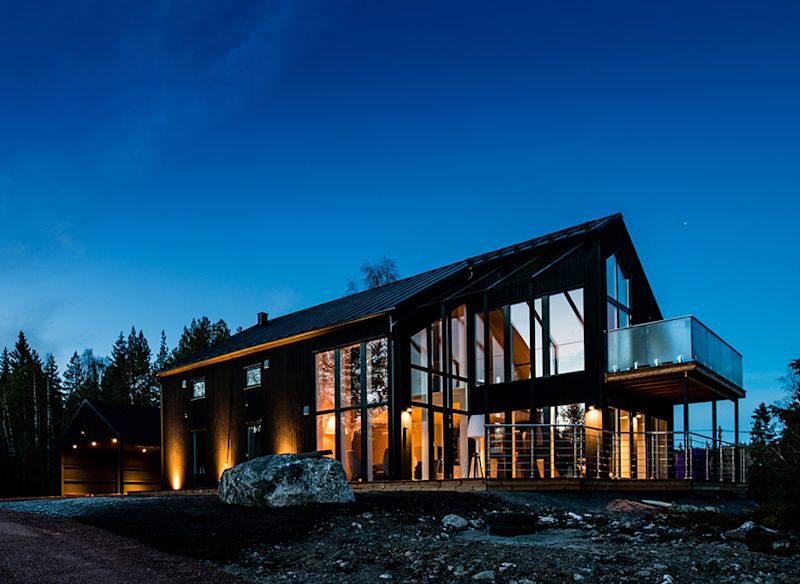 Modern tvåvåningsvilla med glasfasad och varma ljus mot blå kvällshimmel i skogsmiljö.