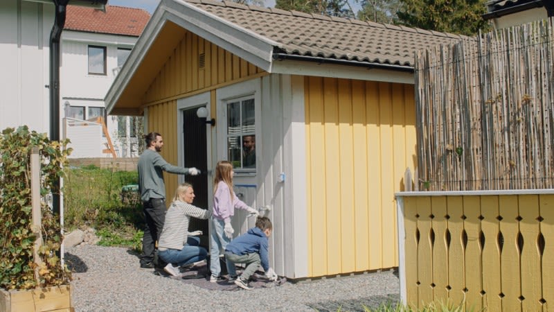 Pappa och tre barn målar en gulvit lekstuga på grusgång i trädgård.