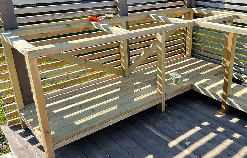 Delvis byggd utebänk i ljus furu på trädäck, spaljéräcke i bakgrunden och verktyg.