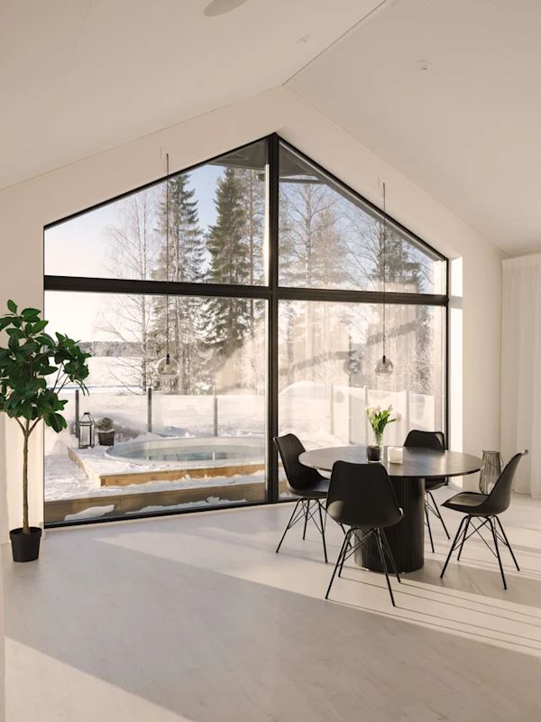 Ljust vardagsrum med svart runt bord och stolar vid stora panoramafönster mot snötäckt skog