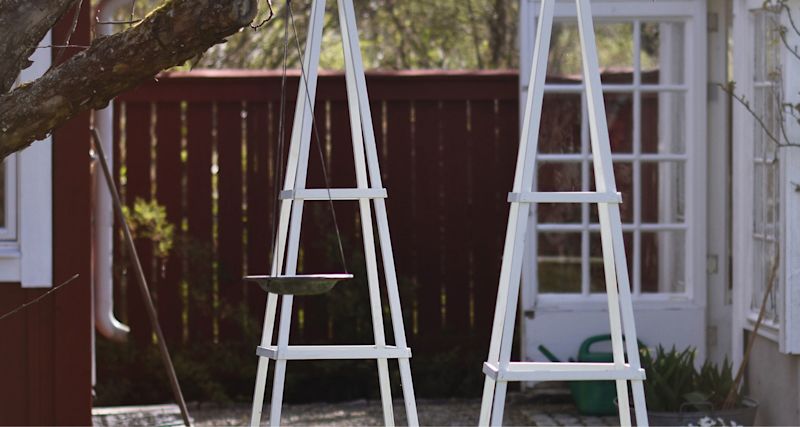 Två vita växtstöd med hängande skål framför röd fasad och vit pardörr.