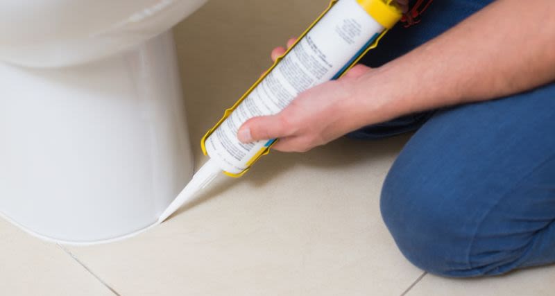 Närbild på person i blå jeans som tätar en vit toalett mot beige klinkergolv med vit fogmassa.