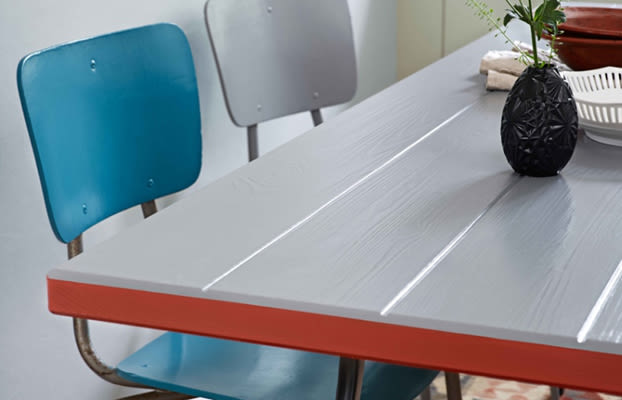 Grått bord med röd kant, två stolar (turkos och ljusgrå), svart vas med grönt blad.