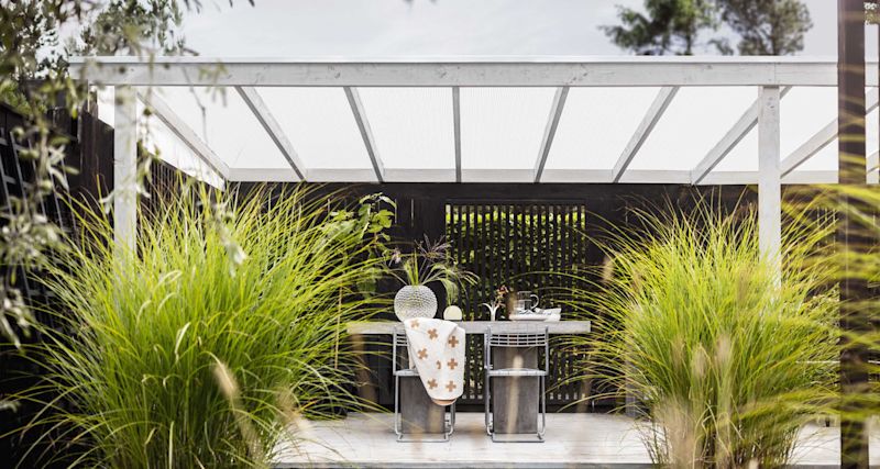 Uteplats med vitt pergolatak, betongbord och stålstolar, omgiven av frodigt grönt prydnadsgräs