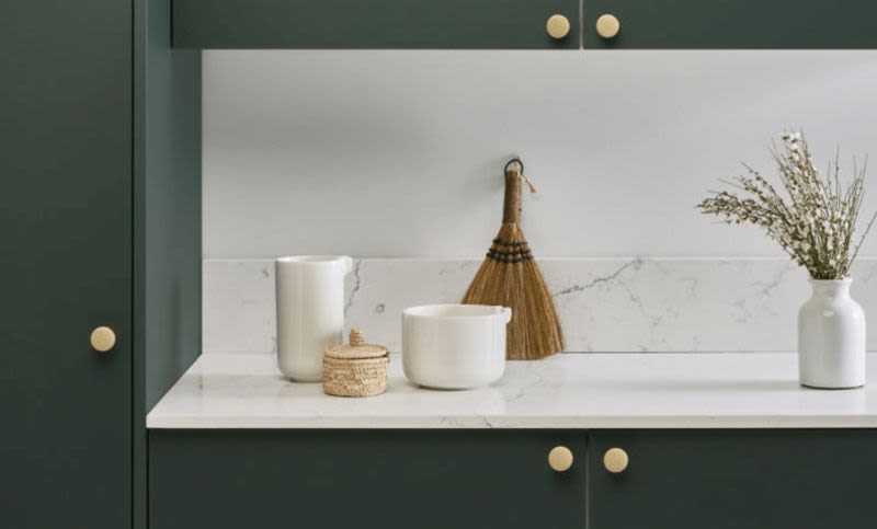 Mörkgröna skåp med mässingsknoppar, vit marmorbänk med krus, borste, vas med torkade blommor