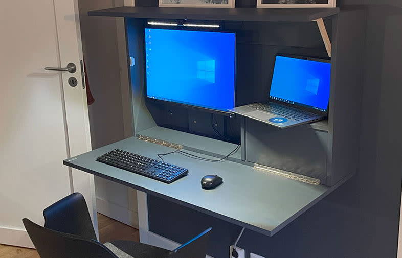 Väggmonterat mörkgrått fällbart bord med två blå skärmar, tangentbord, mus och svart stol