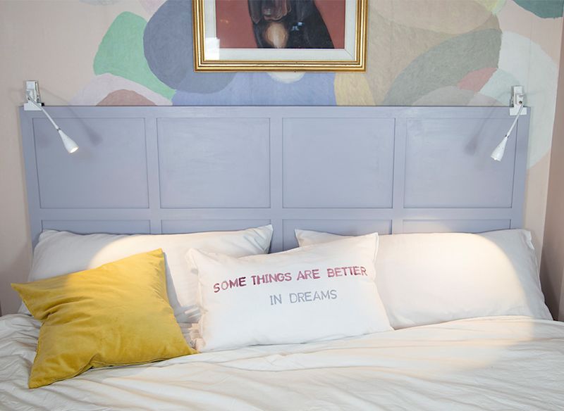 Säng med ljuslila gavel, gul kudde och vit kudde med rosa/blå text samt två vägglampor