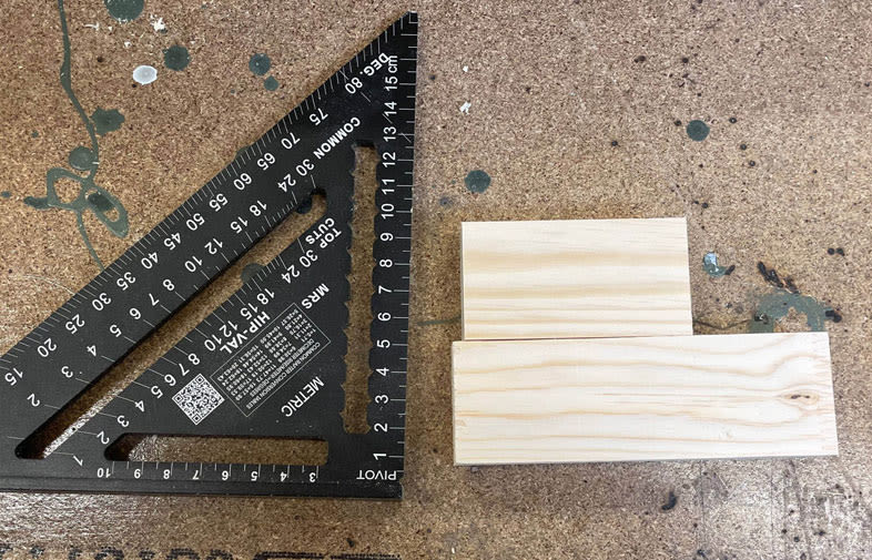 Svart triangelsmått med vita markeringar vid två ljusa träbitar på färgstänkt arbetsbänk