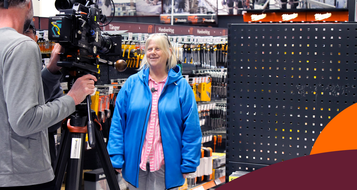 TV-besättning filmar person i blå jacka framför en hylla med gula verktyg i järnhandeln.