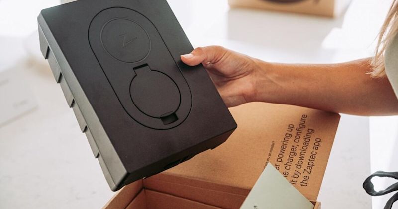 Hand håller en svart rektangulär laddare med två cirkulära lock ur en brun kartong