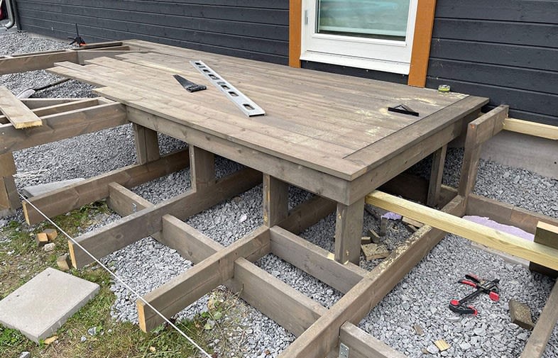 Delvis uppbyggd brun träterrass vid svart husfasad med vit dörr, grus och verktyg.