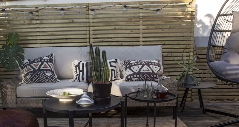 Uteplats med beige rottingsoffa, svartvita mönstrade kuddar, kaktus på svart bord framför träspaljé.