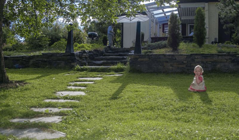 Barn i rosa klänning sitter på grön gräsmatta med stenbelagd gång mot hus med vit parasoll