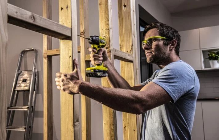 Man i blå t-shirt borrar med gulsvart borrmaskin i ljusa träreglar vid renovering