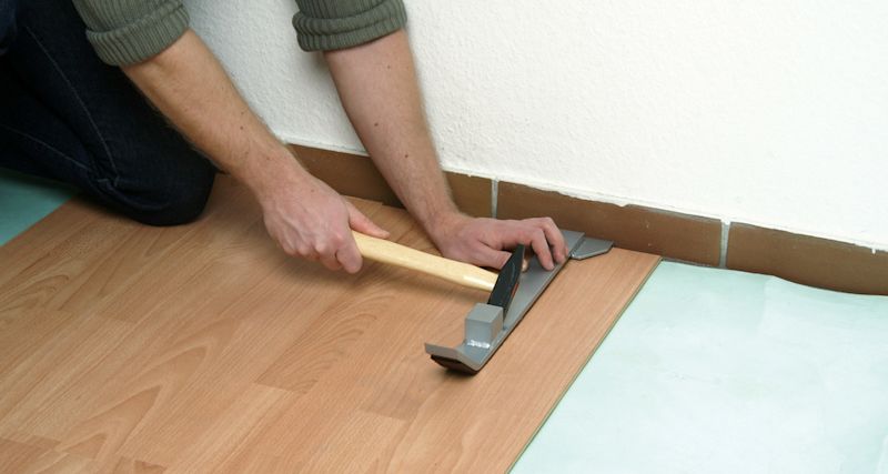 Händer slår fast ljus laminatplanka med hammare och slagkloss vid vit vägg och brun sockel.