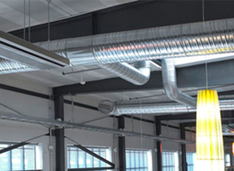 Exponerat industriloft med silvriga ventilationsrör, svarta balkar och gul pendellampa