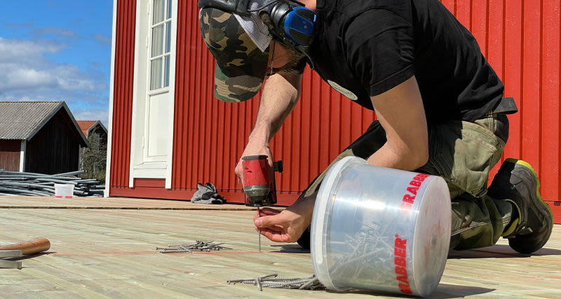 Man med kamouflagemössa och hörselskydd skruvar på ljus trätrall framför rött hus.