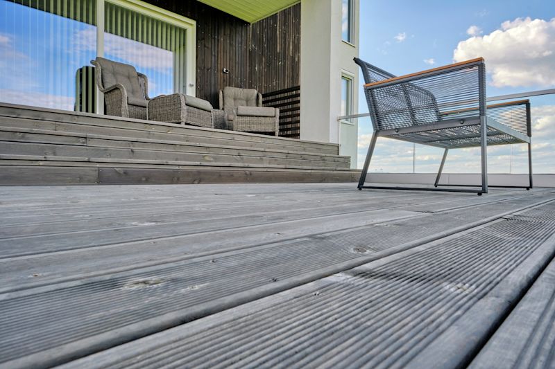 Grå träaltan med flätade fåtöljer, grå metallbänk och glasräcke mot blå himmel.