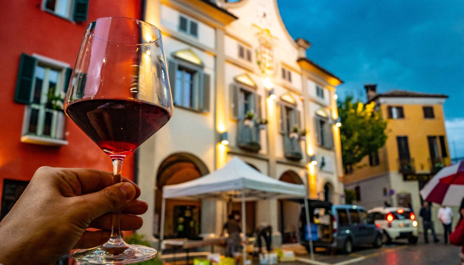 A hand holding a glass of red wine with an Italian town in the background - Barolo