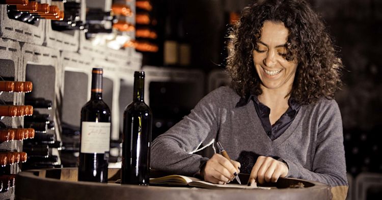 A female winemaker with dark curly hair sitting by a large barrel. There are wine bottles on the barrel and in the background. She is smiling and writing notes on a pad.