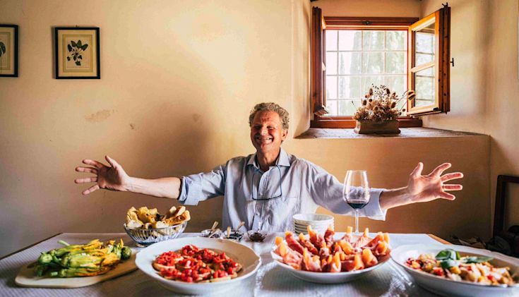 Italian man behind a table with wine and food welcoming with open arms