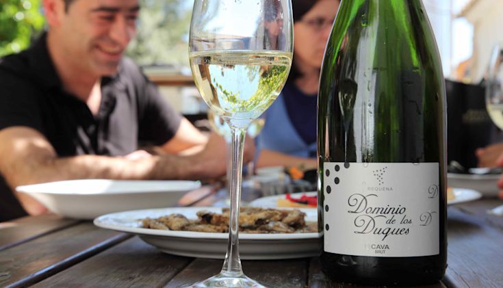 people sitting outdoors at a table with food, with a bottle and glass of Cava in the foreground