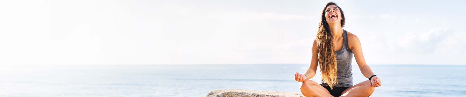 Eine Frau mit langen Haaren sitzt im Schneidersitz am Strand und lacht fröhlich mit dem Meer im Hintergrund.