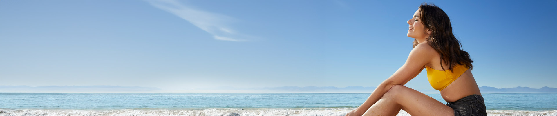 Frau in gelbem Bikinioberteil und Shorts sitzt am Strand, die Augen geschlossen und lächelnd