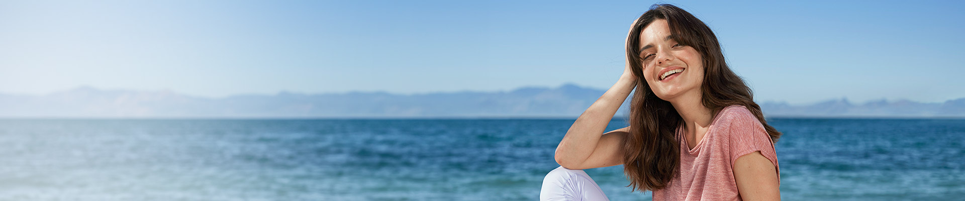 Eine Frau sitzt lächelnd am Strand und trägt ein rosa Hemd und eine weiße Hose mit Wasser und Bergen im Hintergrund.