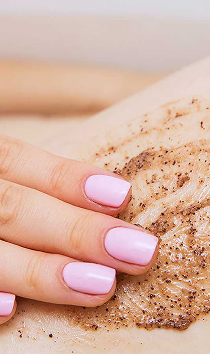Eine Nahaufnahme einer Hand mit rosa Nägeln, die ein Körperpeeling auf die Haut aufträgt und eine glatte, exfolierte Textur zeigt.