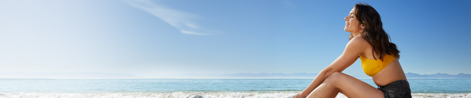 Eine Frau mit langen braunen Haaren lächelt, während sie in einem gelben Top am Strand sitzt, mit dem Meer und dem Himmel im Hintergrund.