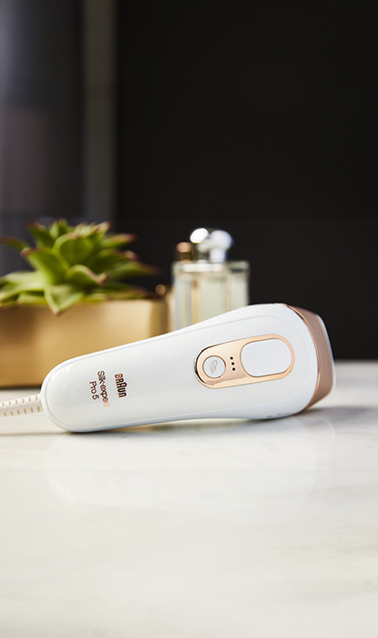 Braun IPL-Gerät mit weiß-goldenem Design auf einer Marmorarbeitsplatte, im Hintergrund Pflanzen und Schönheitsprodukte.