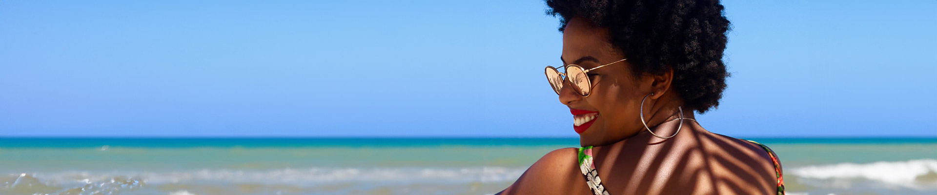 Eine Frau mit lockigem Haar und Sonnenbrille lächelt, während sie am Strand sitzt und das Meer im Hintergrund hat.