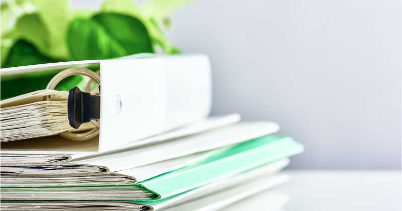 Ein Stapel Ordner und Mappen auf einem Schreibtisch mit sichtbaren Blättern, wobei der Schwerpunkt auf effizienter Organisation mit einer sanften Farbpalette aus Grün und Weiß liegt.