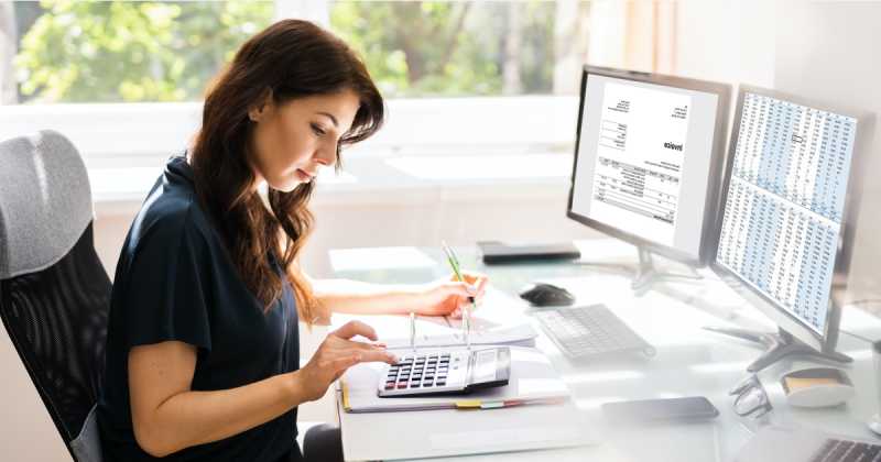 Eine Frau konzentriert sich auf Berechnungen mit einem Tischrechner an einem Schreibtisch mit zwei Monitoren, auf denen Diagramme und Daten angezeigt werden.