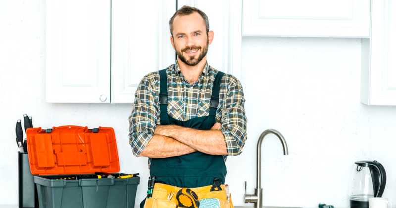 Ein lächelnder Mann mit kariertem Hemd und Werkzeuggürtel steht mit verschränkten Armen in einer Küche neben einem orangefarbenen Werkzeugkasten.