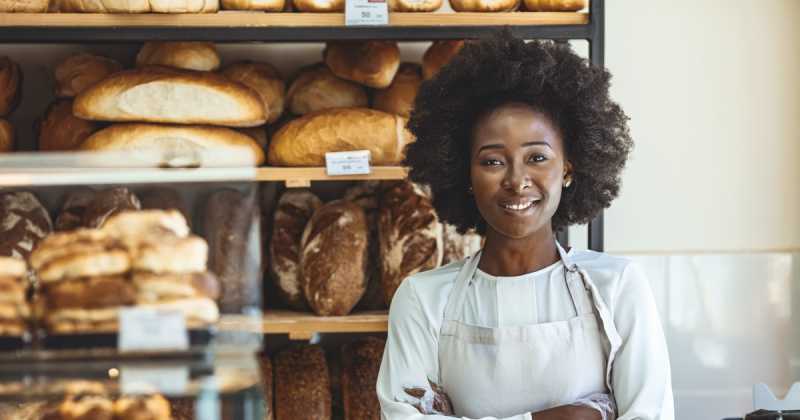 Eine lächelnde Frau in einer Schürze steht vor einem Bäckereiregal, das mit verschiedenen Brotlaiben gefüllt ist.
