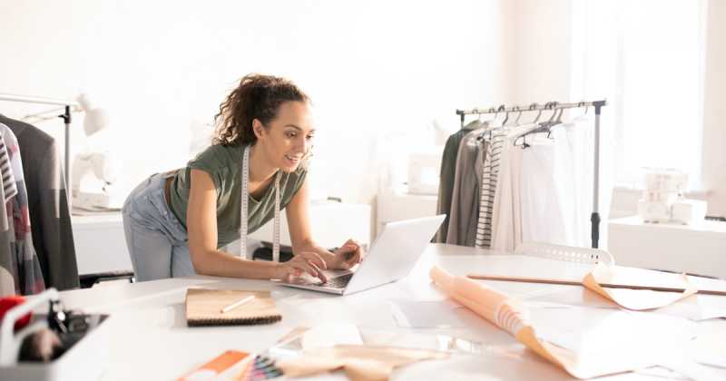 Eine junge Frau arbeitet an einem Laptop in einem hellen Modedesignstudio, umgeben von Stoffmustern und Skizzen.
