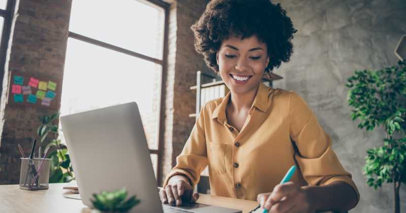 Eine Frau mit lockigem Haar lächelt, während sie an einem Laptop arbeitet und sich in einem modernen Büro mit Haftnotizen im Hintergrund Notizen macht.