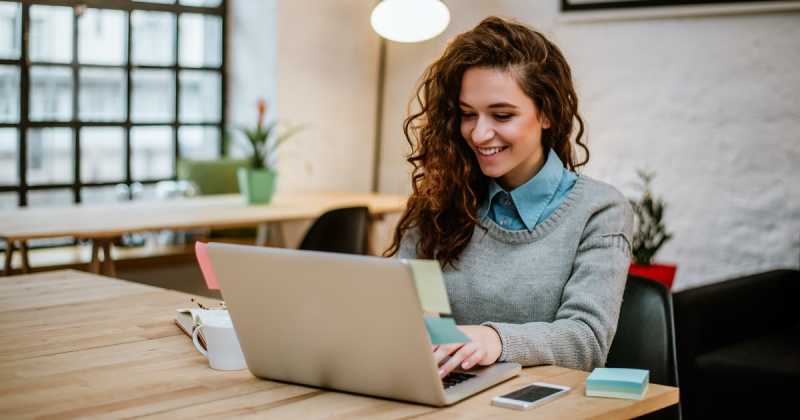 Eine junge Frau lächelt, während sie an einem Holztisch in einem modernen Büroumfeld an einem Laptop arbeitet.
