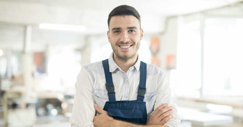 Ein junger Mann in blauer Schürze und weißem Hemd lächelt selbstbewusst in einem hellen Arbeitsbereich.
