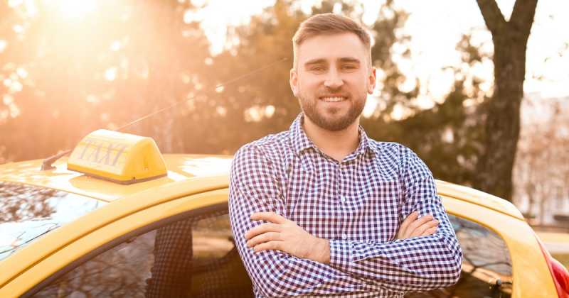 Lächelnder Mann mit verschränkten Armen steht neben einem gelben Taxi in einer sonnigen Parklandschaft.