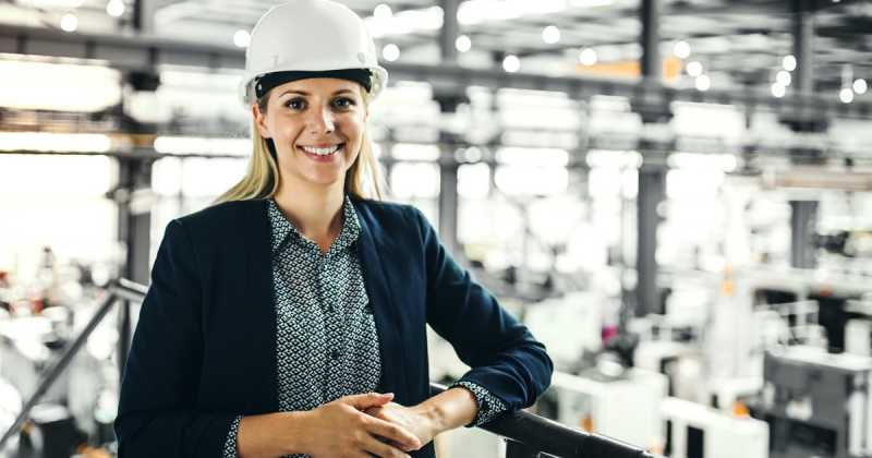 Eine lächelnde Frau mit Schutzhelm und Geschäftskleidung steht in einer Industrieanlage.