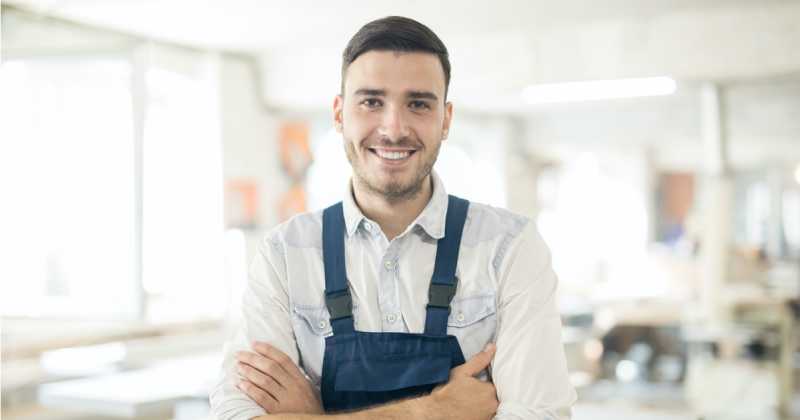 Ein lächelnder Mann in weißem Hemd und blauem Overall steht mit verschränkten Armen in einem hellen, industriellen Arbeitsbereich.