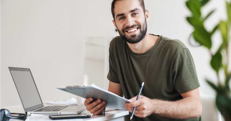 Ein lächelnder Mann mit Bart hält ein Klemmbrett und einen Stift in einem gut beleuchteten Heimbüro, in der Nähe stehen ein Laptop und ein Smartphone.