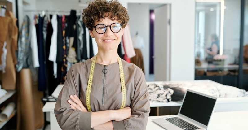Eine selbstbewusste Modedesignerin mit lockigem Haar und Brille steht mit verschränkten Armen in einem Studio, im Hintergrund sind Kleidung und ein Laptop zu sehen.