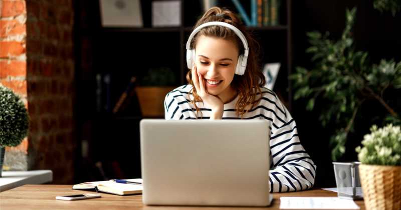 Eine junge Frau mit Kopfhörern lächelt, während sie an einem Schreibtisch mit Büchern und Pflanzen einen Laptop benutzt.