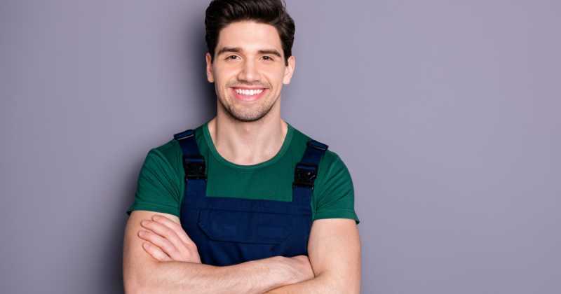 Ein lächelnder Mann in grünem Hemd und blauem Overall steht mit verschränkten Armen vor einem grauen Hintergrund.